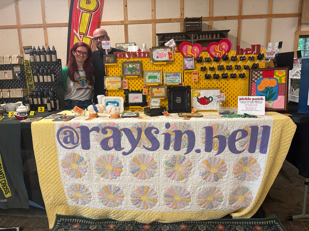 A booth table at a craft fair with a quilted yellow and white tablecloth featuring the text '@raysin.hell' in large purple letters.