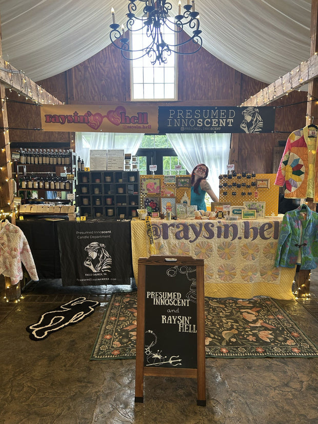 A rustic indoor market or booth display with a woman standing behind a table under draped white fabric and a chandelier. Multiple signs read 'Presumed Innocent' and 'Raysin Hell,' with shelves of packaged products, hanging garments, and decorative textiles around the space.